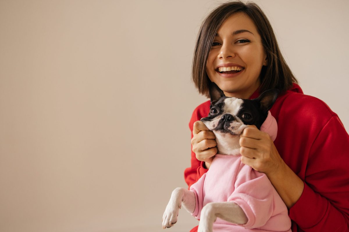 Smiling young caucasian woman holding her pet's cheeks in hoodie on white background. Love between dog and owner concept